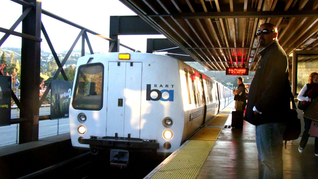 the front of a BART train stopped at a station with a passenger waiting on the platform