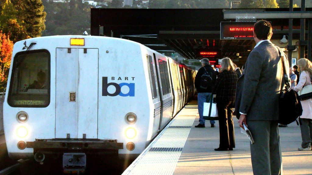 A subway train is stopped on the left side of the frame. On the right, dozens of people, only their backs visible, stand on a station platform, outside, waiting to board.