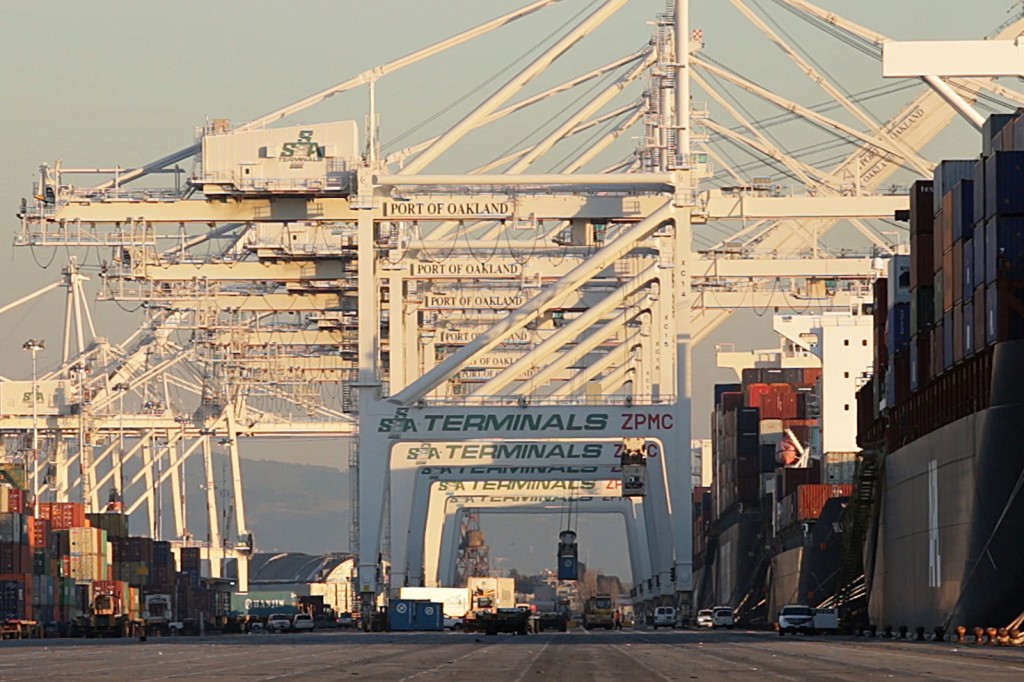 A terminal at the Port of Oakland shows a series of elevated horizontal white steel bars with the words "Port of Oakland" over frames that say "Terminals ZPMC"