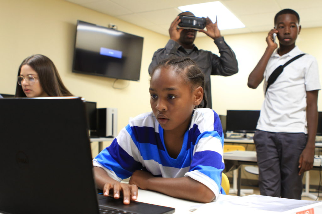 Xavier Stroud, 10, of East Oakland uses 3D technology gaming software at the David E. Glover Education and Technology Center. Youth Program Director, Paul Williams, exhibits a VR game in the background. Credit: Stefanie Le.