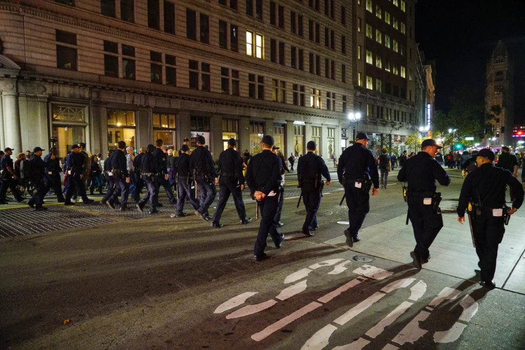On city street at night, by the words painted STOP in the street, a wall of uniformed police officers walk forward. 