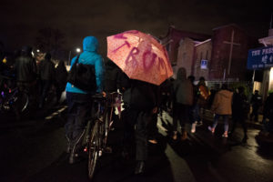 Although rain was forecast, the evening march was mostly dry. Here, marchers walk along Broadway under an umbrella-turned-protest sign. Photo by Rosa Furneaux.