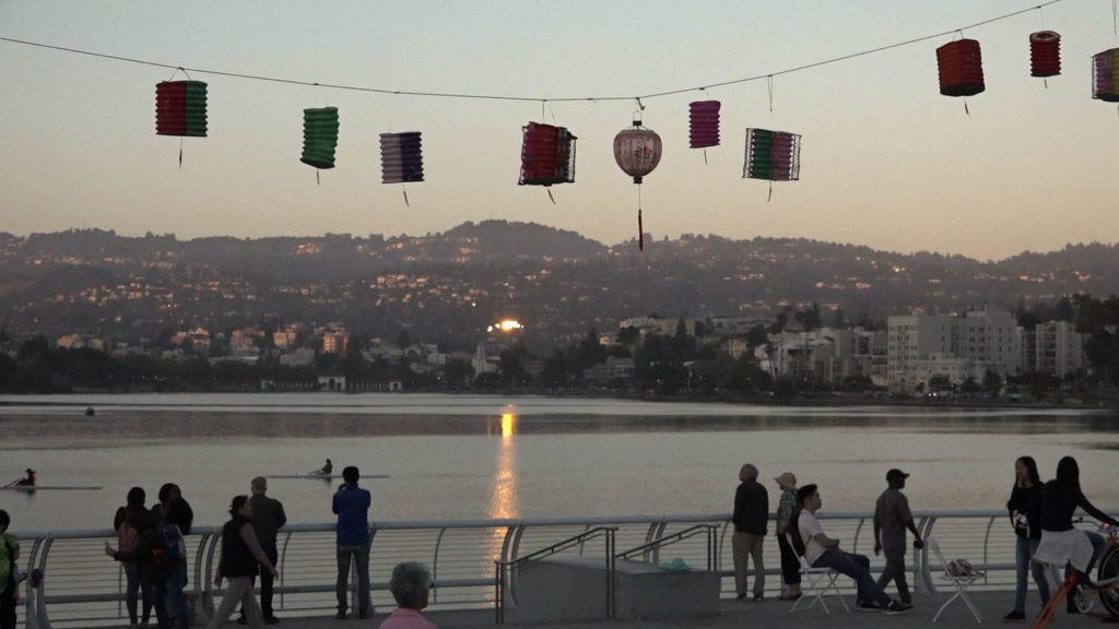 Ceremony attendees and passersby watch lanterns float along Lake Merritt near sunset.