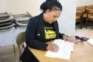 Mayoral candidate Cat Brooks sits at a table, looking over a ballot, a pen in her right hand, which has a ring on it. She is wearing a blue and yellow Brooks for Mayor T-shirt under a blue blazer. Her braided hair is pulled into a bun and an earring dangles fro her right ear.