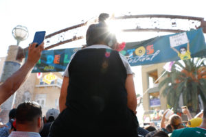 A child sits on an adult’s shoulders to see above the dense crowd during one of the traditional performances by Aztec dancers at the Unity Council’s Día de los Muertos festival on November 4. Just ahead, a sign hangs from a neighborhood arch, with artwork by local artist Eduardo Chaidez.