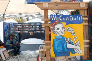 Artist Sean Levon Nash sits inside his stand as customers peruse nearby. Nash, who was a regular at the festival 15 years ago, started going back more regularly the last few years. He sells Día de Los Muertos-themed paintings and jewelry, like a poster that combines skeleton motifs with Rosie the Riveter.