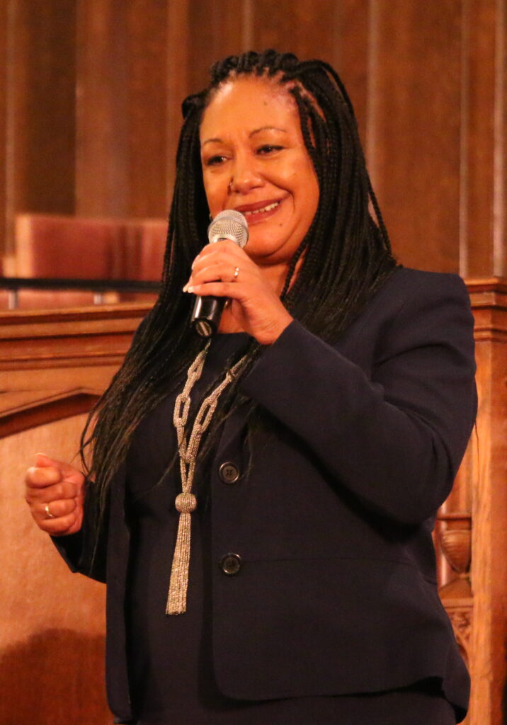 Oakland mayoral candidate Cat Brooks stands in long braided hair, a microphone in her right hand, wearing a braided bold necklace and all black clothing
