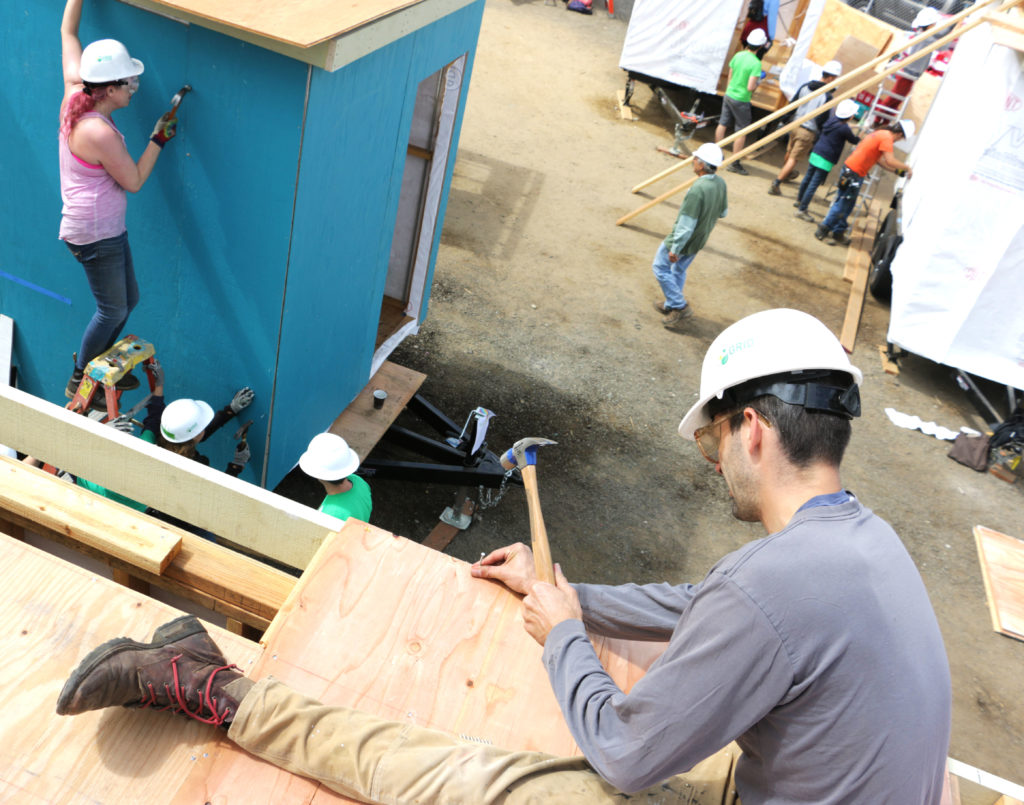 A volunteer in a white hardhat hammer boards on the roof of a tiny house.