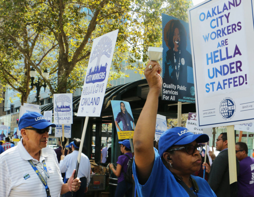 At a protest, a city worker raises a fist in the air while holding a sign that says "Oakland city workers are hella understaffed."