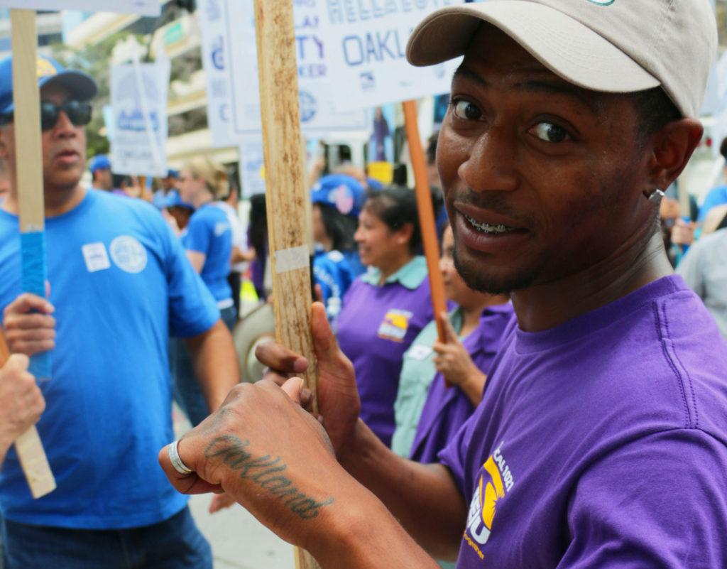 An SEIU member shows off an tattoo that says "Oakland" on their hand. People carrying picket signs walk in the background.