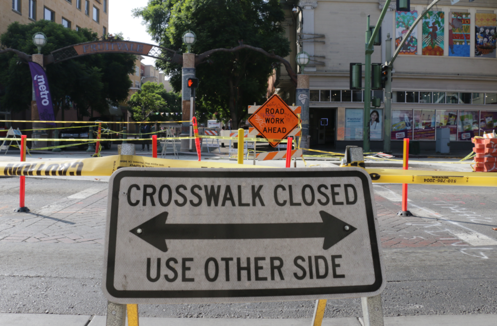A sign says "Crosswalk closed-used other side," and traffic cones cordon off the center of the street on International Boulevard.