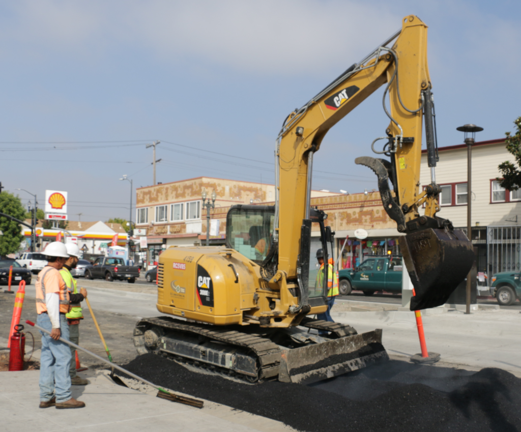 A backhoe pours and moves concrete as construction workers pave