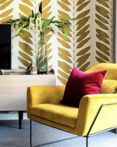 This is a picture of a yellow-themed living room. There is a yellow chair with a pink pillow. The wall has gold leaves on it.