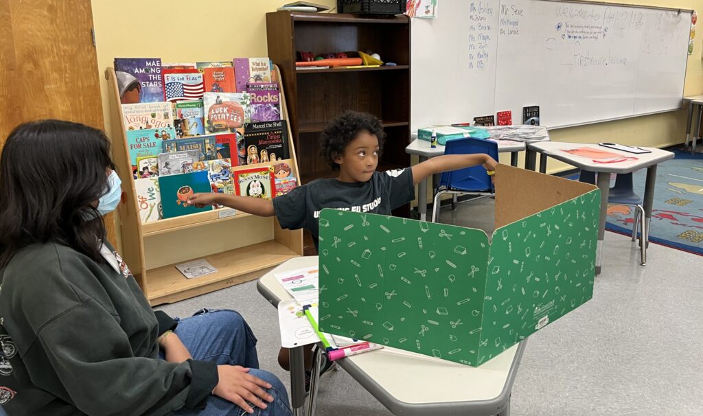 Second grader Miles makes a 180 degree angle with his arms outstretched, facing the camera, while his tutor Briana Ortiz watches, seated in the classroom.