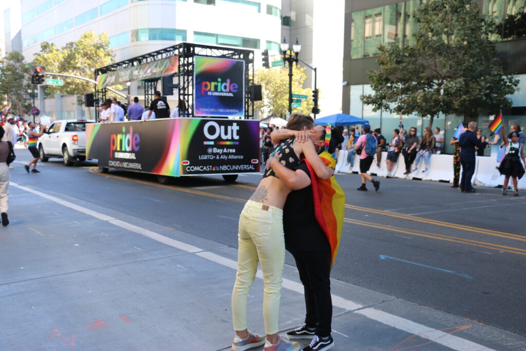Two women hug in the middle of an Oakland street, one draped in a rainbow flag, the other in white jens and a short black top.