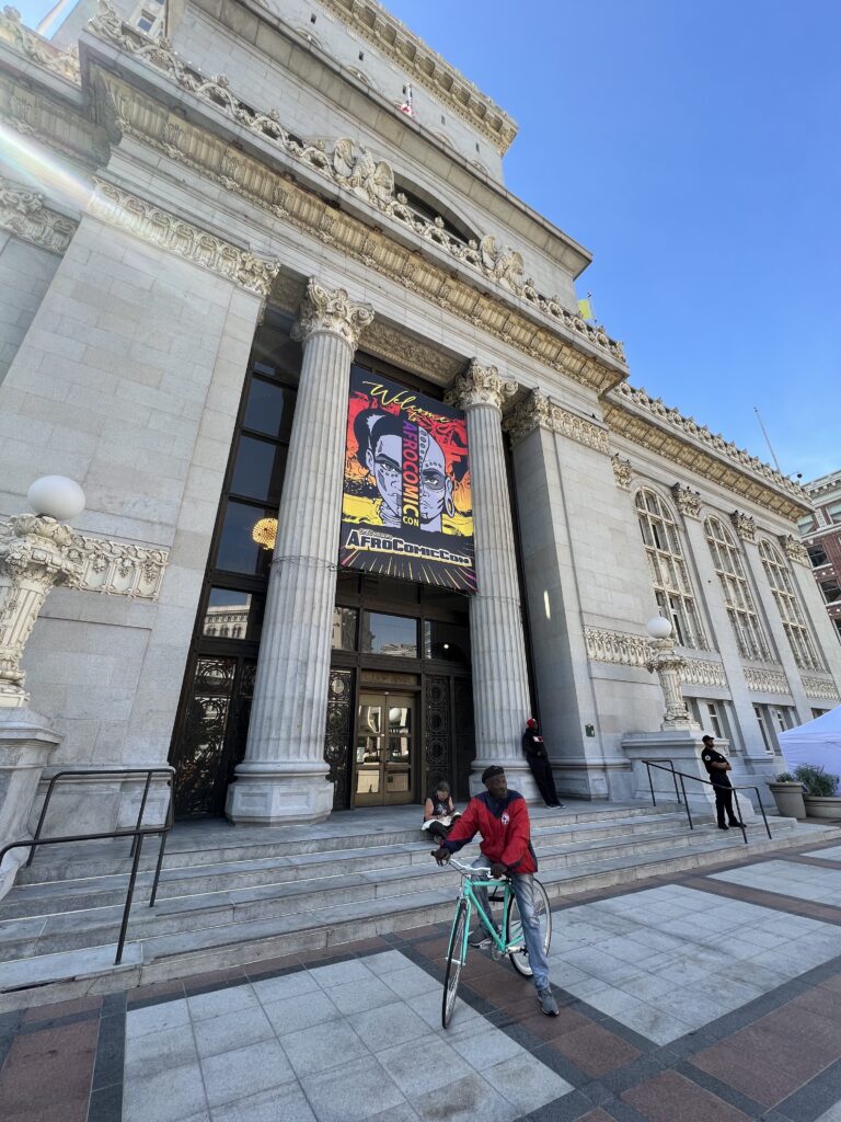 Bicyclist in a red jacket pauses outside Oakland City Hall in front of a AfroComicCon sign.