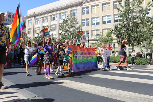 About 15 children waving rainbow flags walk behind an OUSD rainbow banner on an Oakland street.