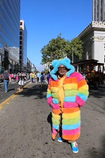 A man stands in the middle of an Oakland street wearing a puffy, full-length rainbow coat.