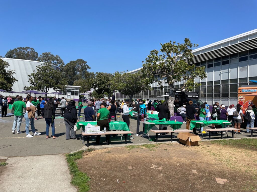 Dozens of people stand around picnic tables topped with green paper tablecloths outside a school.