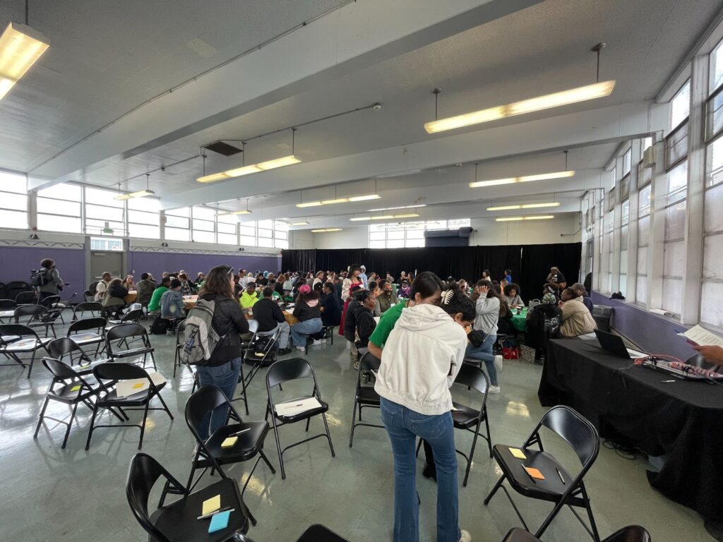 Dozens of people get into groups in black folding chairs in a school auditorium.