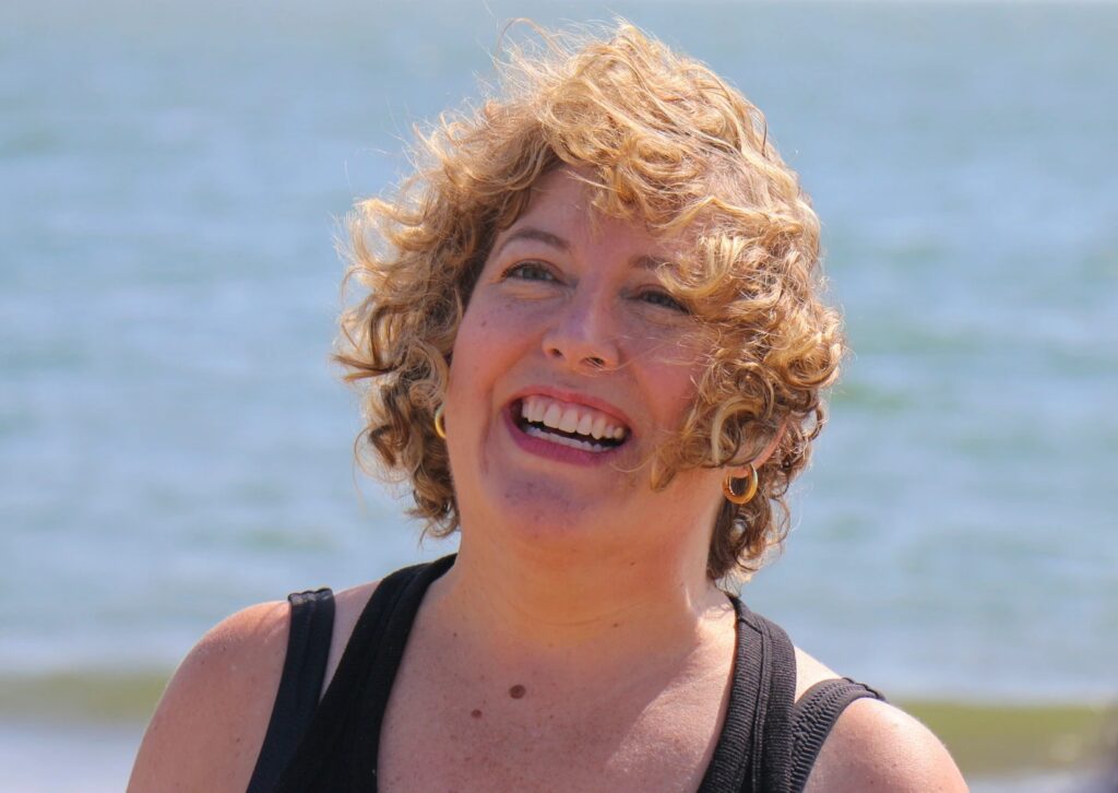 A closeup of a woman's smiling face. She has short sandy blonde hair and is standing with the bay behind her.