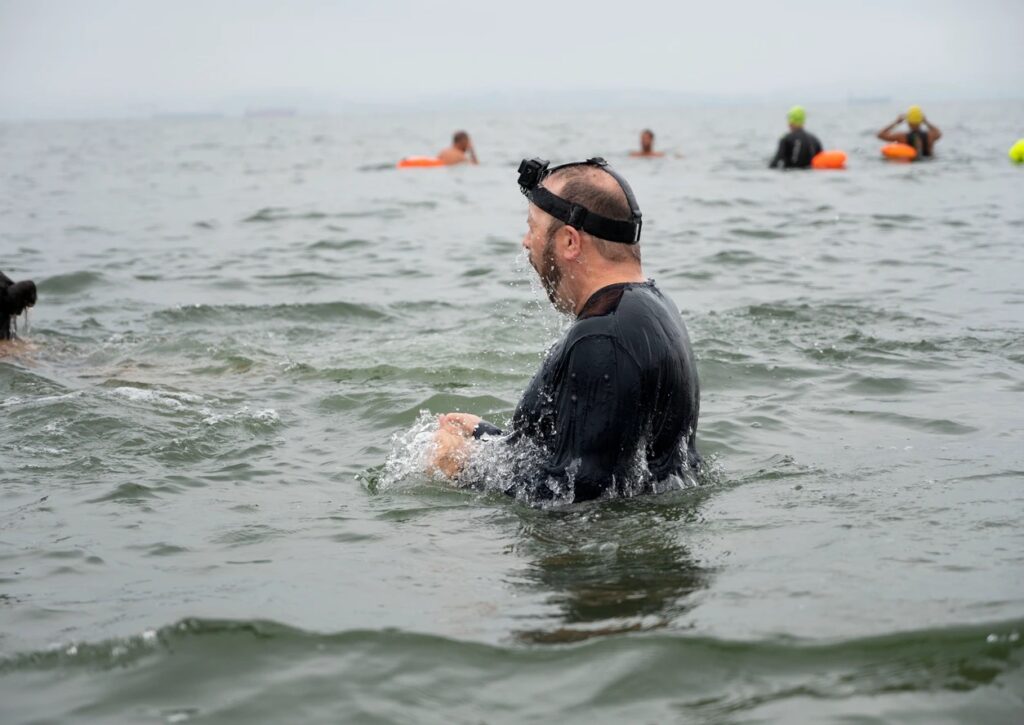 A man in a long-sleeved black shirt is chest high in the bay with his goggles on top of his head. In the blurry background are four other swimmers.