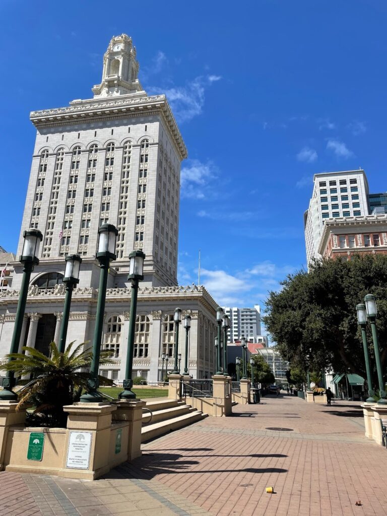 Oakland City Hall is depicted in this photograph, featuring a stately gray building adorned with a prominent clock tower atop its impressive height. The structure is beautifully framed with green streetlights positioned around it.