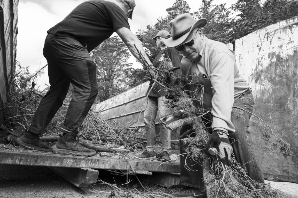 Blacm and white photo of a man in dark clothes and baseball cap on the left, a woman in a long black T-shirt and jeans and cap in the middle and a man in a cowboy hat, jeans and tan jacket all lifting branches onto a cart.