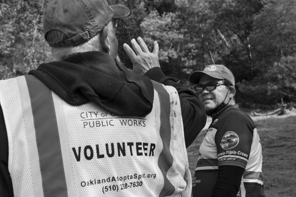 Black and white photo. The back of a man in a cap and vest with the words City of Oakland Public Works Volunteer printed on the back stands in a park talkng to a man to his right who is wearing a cap, glasses and a shirt with the words California Triple Clown on the left sleeve.