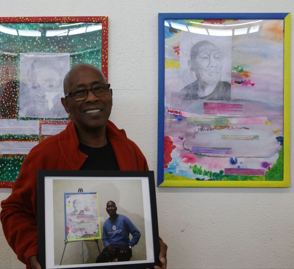 Keith stands in front of two paintings, holding a photo of himself with one of his artworks. He wears a red sweater over a black T-shirt, black glasses, and smiles at the camera.