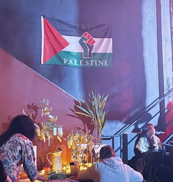 A Palestinian flag, red triangle turned sideways on the left, then a black, white and green stripe fille the rest, with a fist in the middle hangs above an altar that had a few candiles on it and photos.