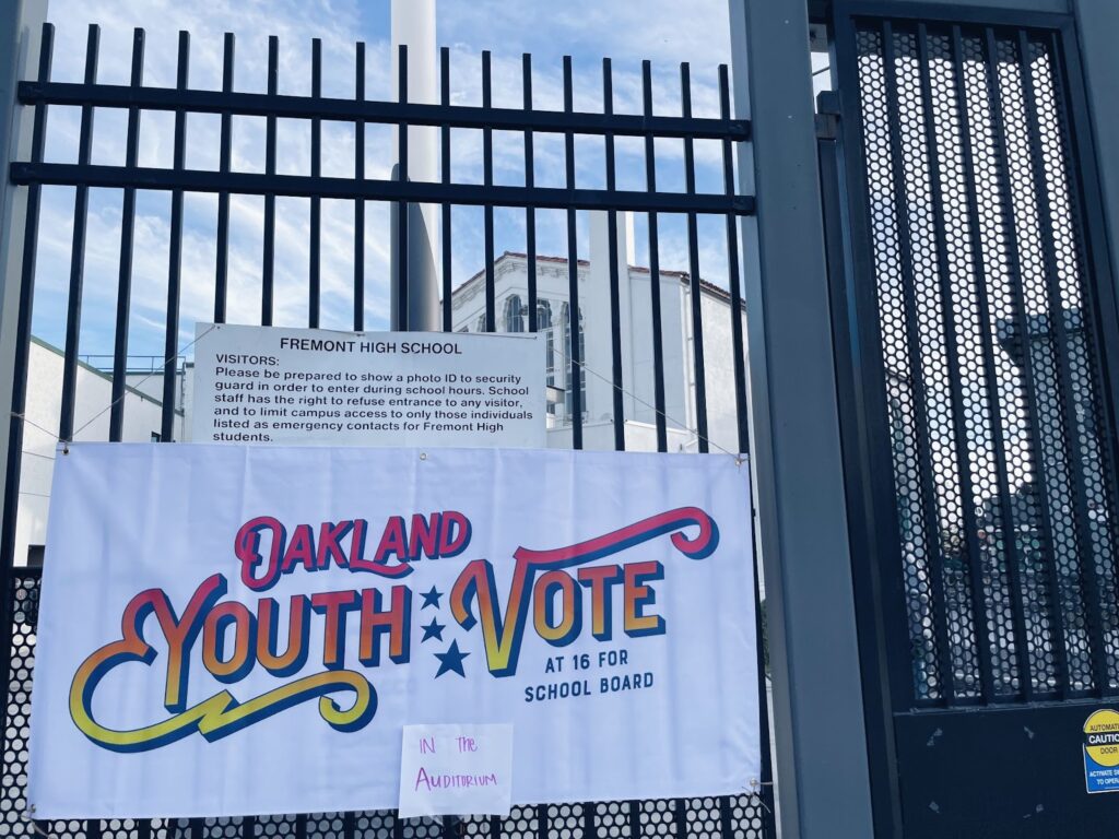 A white banner with red, orange and yellow lettering hangs on the wrought iron gate leading to Fremont High says: Oakland Youth (three vertical stars) Vote at 16 for school board. Taped to that banner is a piece of paper with the words "in the auditorium"
