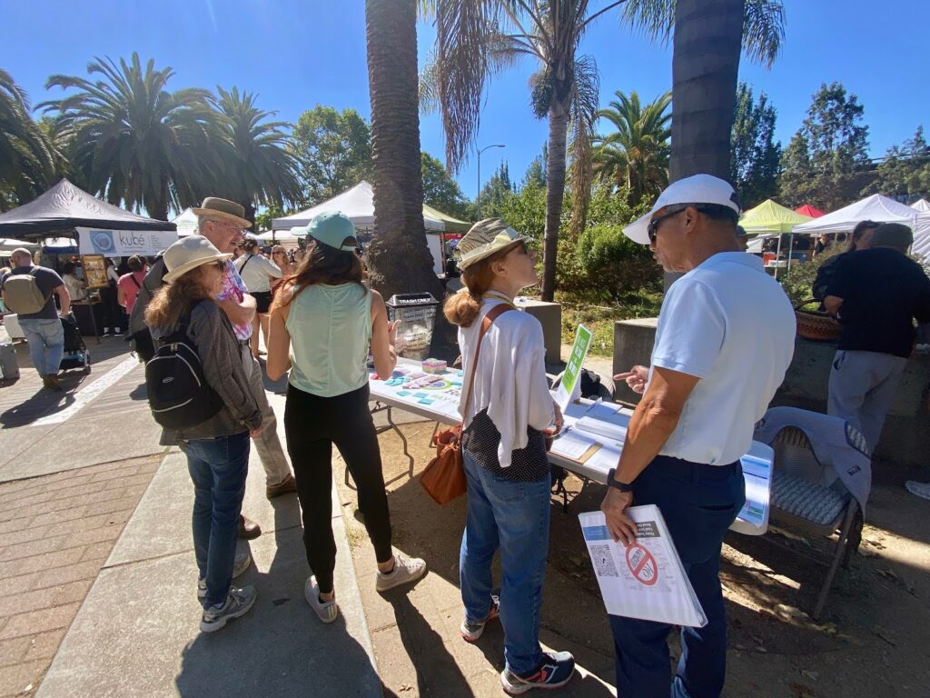 On a sunny day, about a dozen people, five prominent in the foreground, stand around a table chatting with each other, pamphlets in hand. All are in jeans and sneakers and wearing hats or caps.