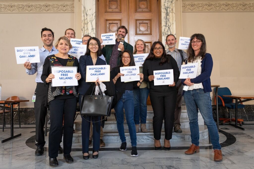 11 people stand in uneven rows, all smiling at the camera, all holding signs that read "Disposable Free Oakland!"