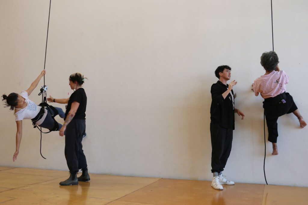 On the far left and on the far right are women in harnesses dangling from ropes, trying to scale a wall. Each is assisted by a teacher all in black.