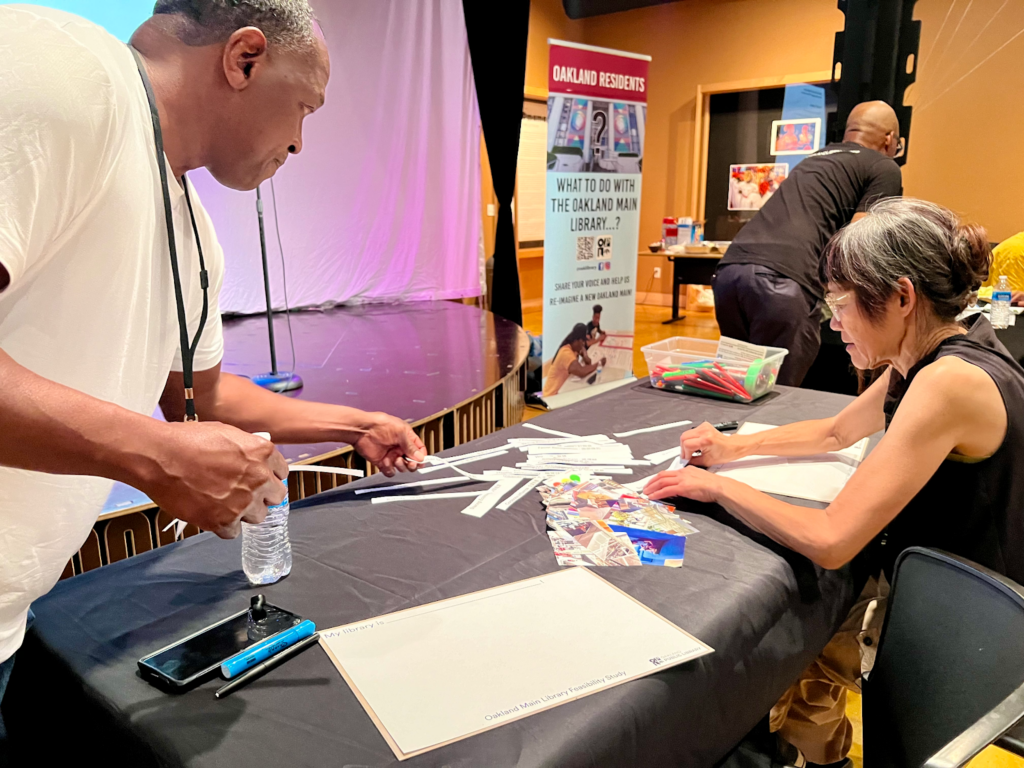 A man stands on the left, his right hand clenches, his left extended with a strip of paper on it over a table with a black cloth and scraps of papers, a box of markers and some paper. A woman is at the table in salt and pepper hair, pasting something on the paper.