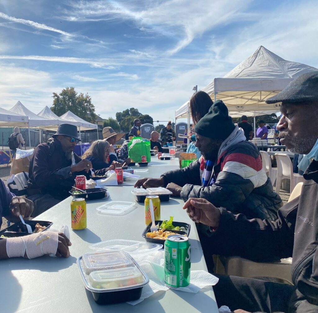 About 10 people are visible around a long table set up outside, with soda cans and trays of food on top. Around them are booths with white canopies.