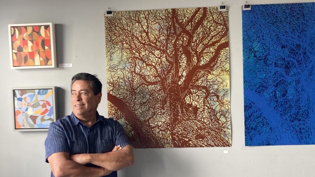 A man in a blue short-sleeved collared shirt stands with arms folded before four prints -- two large and two small, the one closest to him is large and is of tangled tree branches.