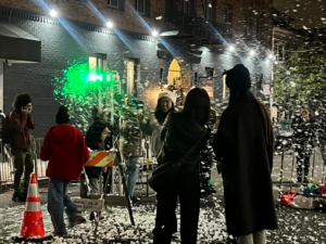 About have a dozen people, some in movement and blurred are pictured outside, in warm clothes, some in hats, with fake snow swirling and on the ground.