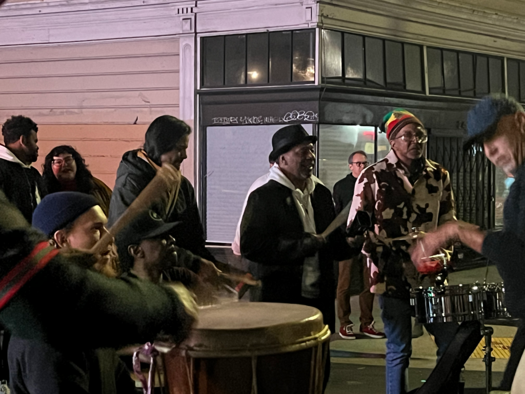 An coat-covered arm is in the left of the frame, banging on a large drum. On the far right, a man in a cap plays a metal drum and cymbal set. Moving around them are about eight people, all in winter coats, some in hats, all men except or one woman.