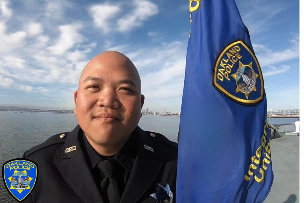 Head and shoulders shot of an Oakland police officer in uniform standing to the left of a blue flad with Oakalnd Police badge on it. The shot is taken with the bay behind the officer, who is smiling, though showing no teeth. He is Asian and bald.