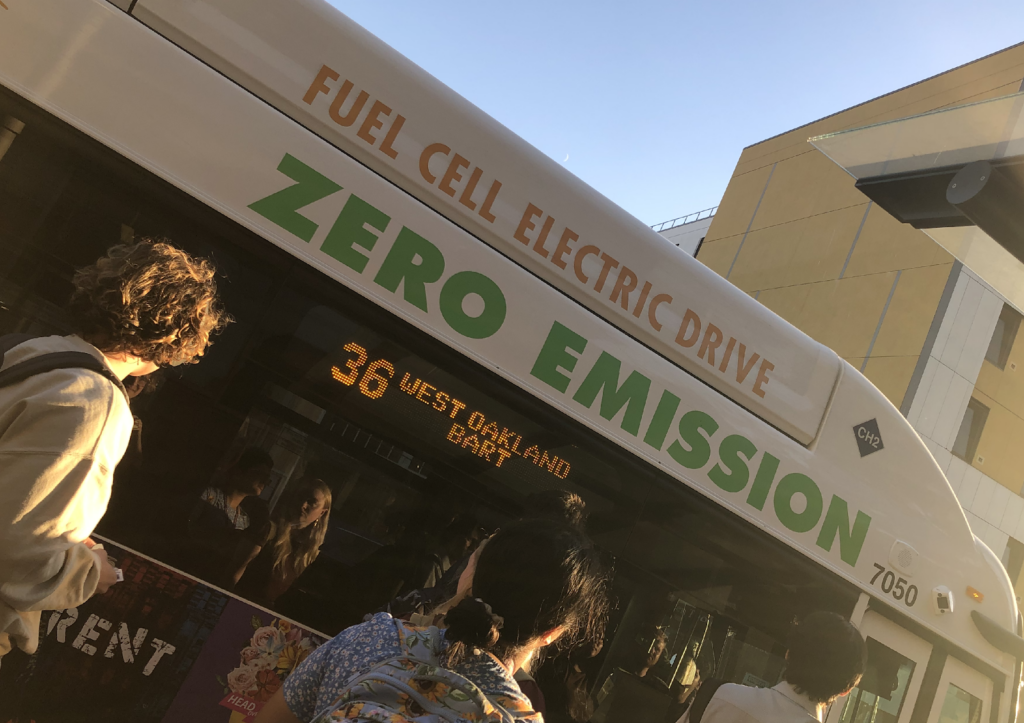 People await to board the AC Transit route 36 bus to the West Oakland BART station. The bus has the words "ZERO EMISSION" in green below the words fuel cell electric drive .