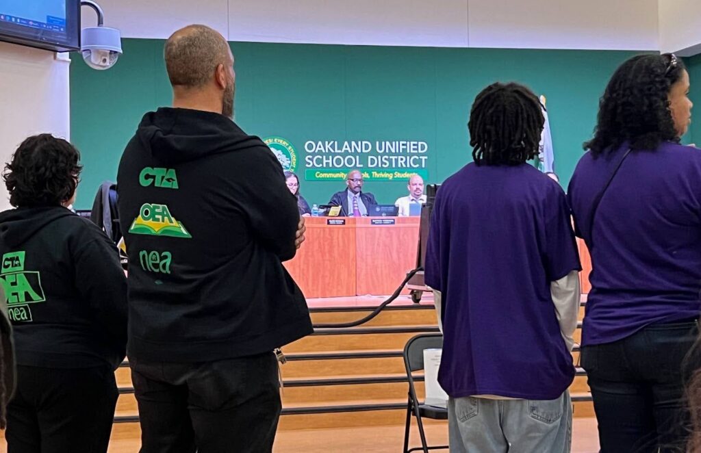 Members of the Oakland Unified School District board sit behind a podium, as teachers union members in black jackets and students in purple T-shirts wait to speak.