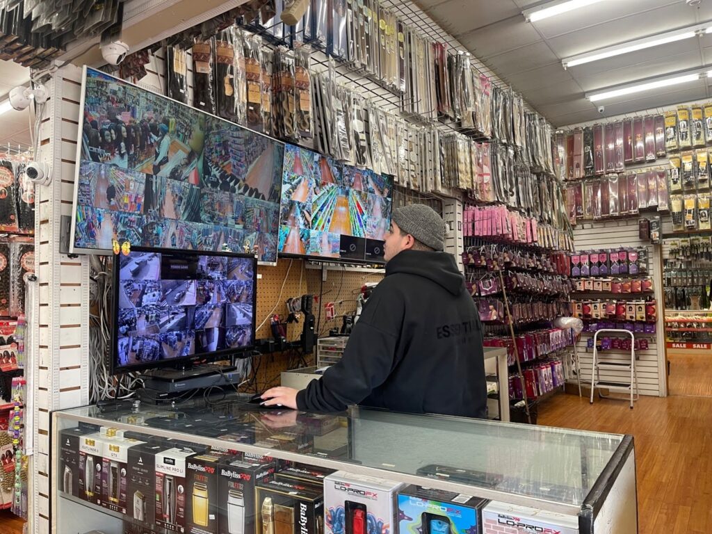 A man in a black hoodie and gray knit cap stands before three screens with surveillance footage, behind a counter that has boxes of hair clippers. On the walls hang lots of packages.