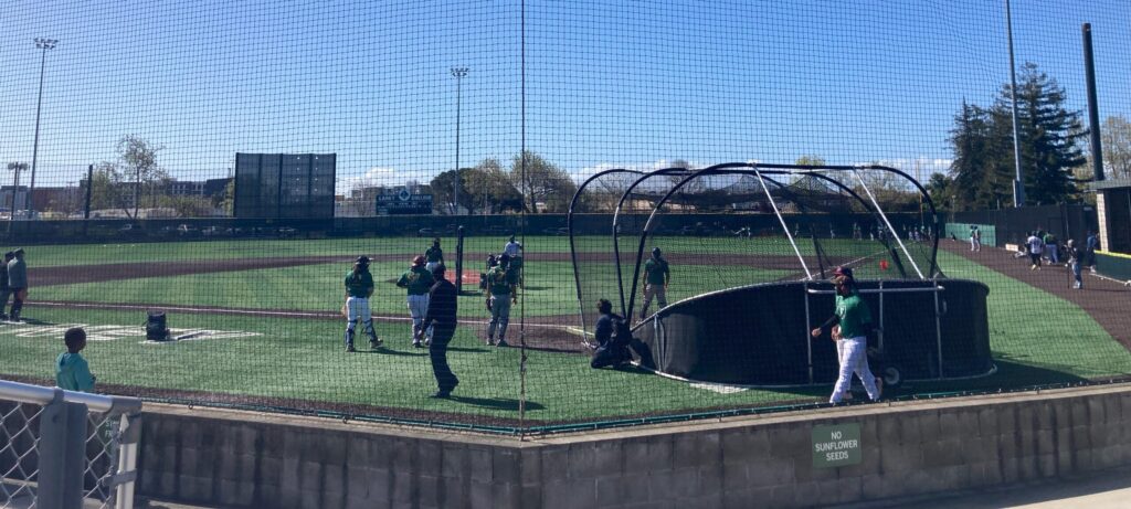 About 10 baseball players mill about, waiting to hit on a baseball diamond that ends with a low fence beyond which are some industrial type buildings.