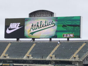 Atop a row of bleachers is a big scoreboard showing Nike and the Athletics, with the time: 12:18 p.m.