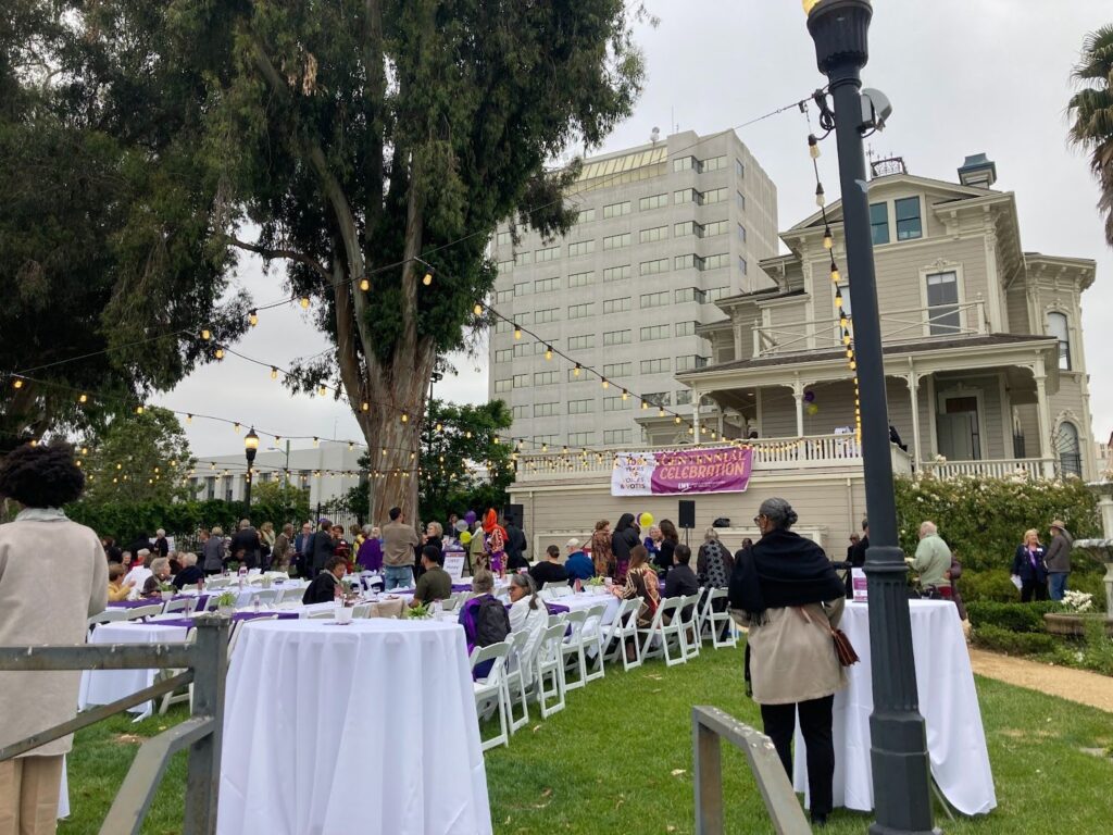 Well-dressed people sit around long white-clothed tables with white chairs on a lawn, under a eucalyptus tree.