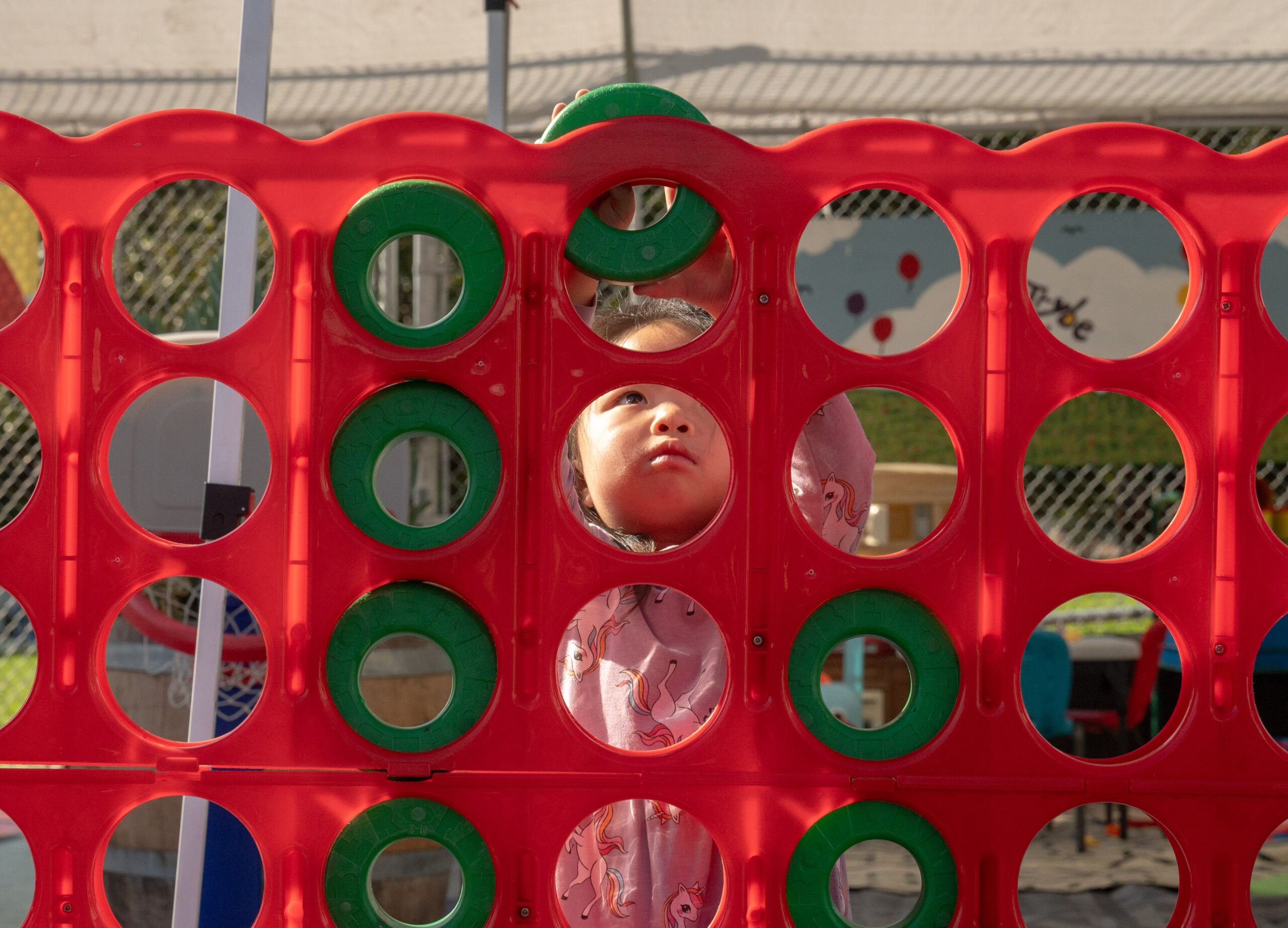 A child's face is visible through a hole in a giant red Connect 4 board.