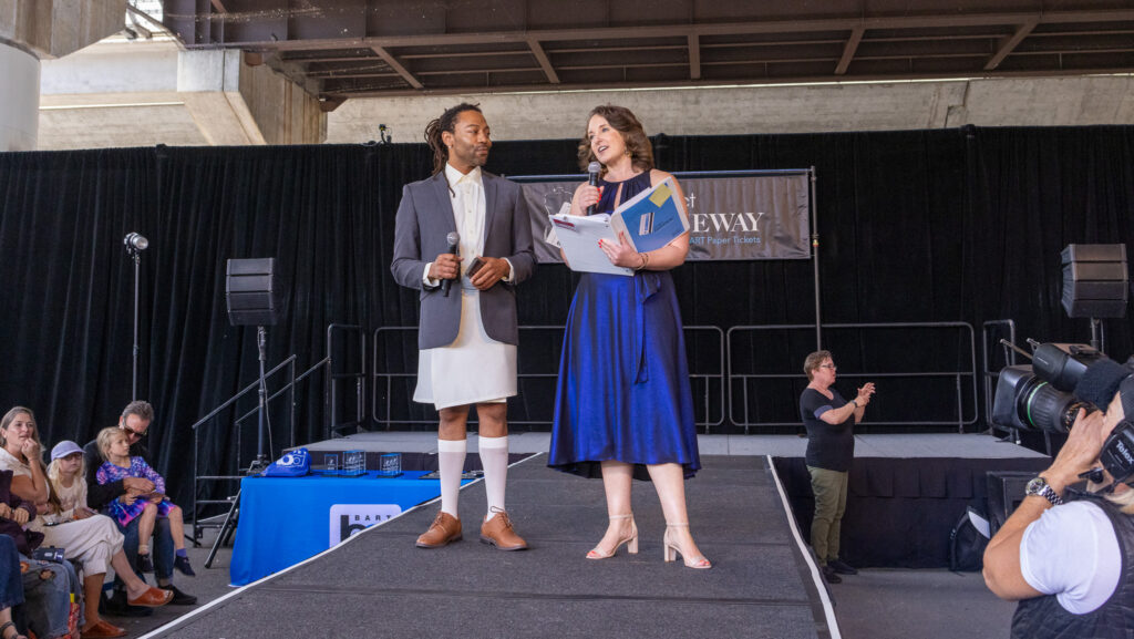 A man in braids and gray suit coat over a white knee-length dress is on the left, being interviewed by a woman on the right with a long blue dress and a binder open in her left hand, a microphone in her right. They are standing on a runway with a few people visible in chairs on the floor.