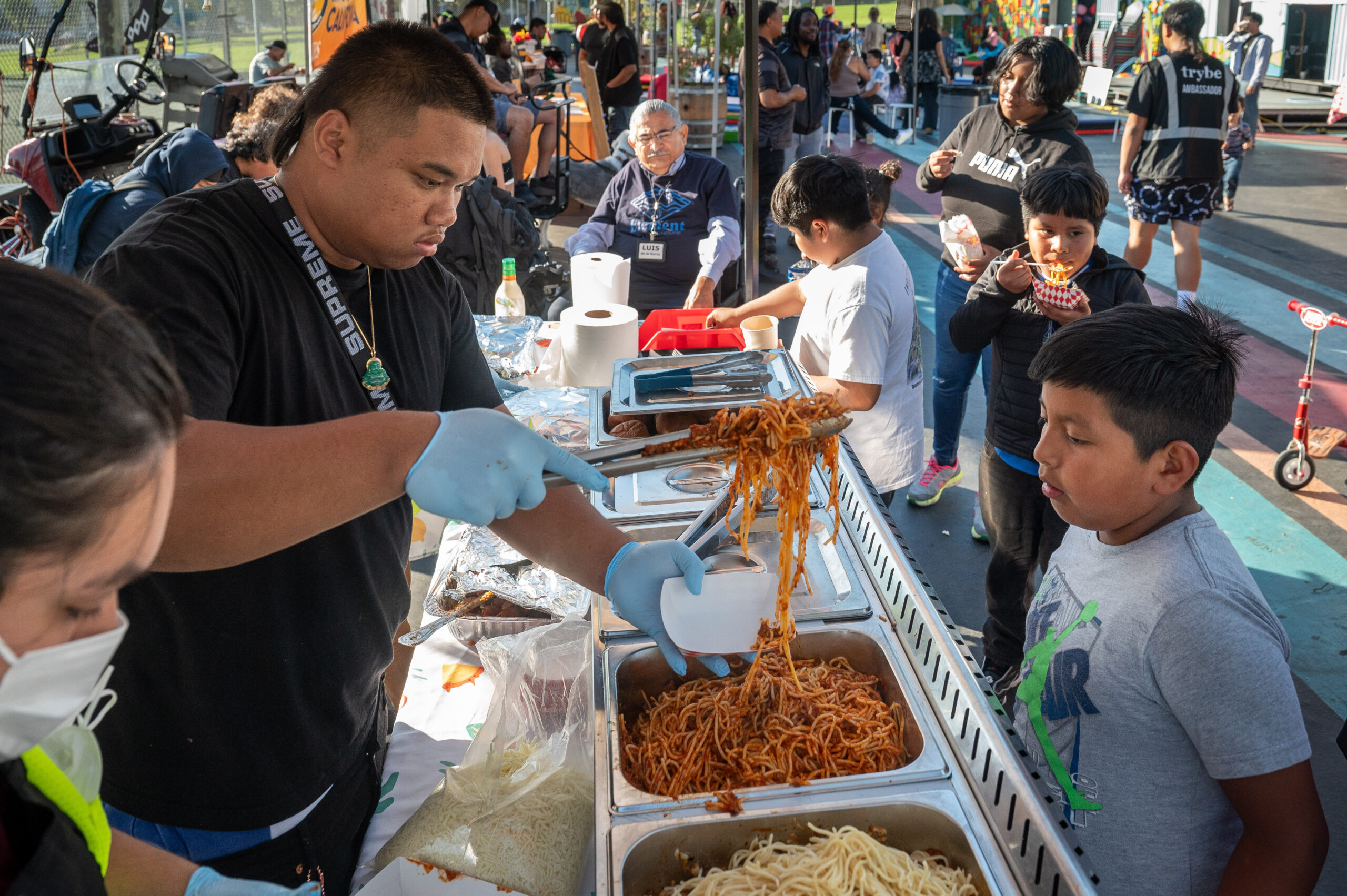 A man serves spaghetti from a big vat to a line of kids.